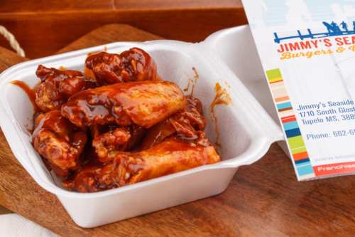 A takeout container filled with saucy chicken wings next to a menu for Jimmy's Seaside Burgers & Seafood.