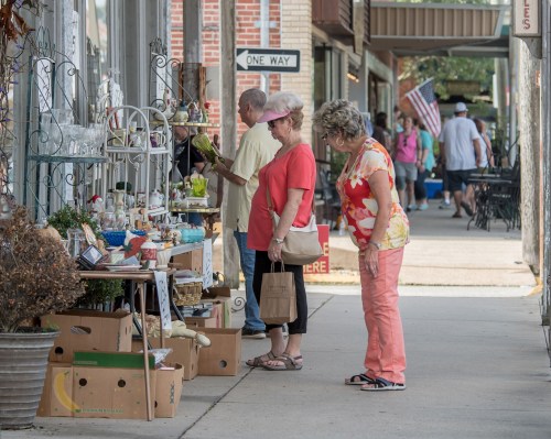Two women browse a display of items outside a shop on a busy street, with people and flags in the background.