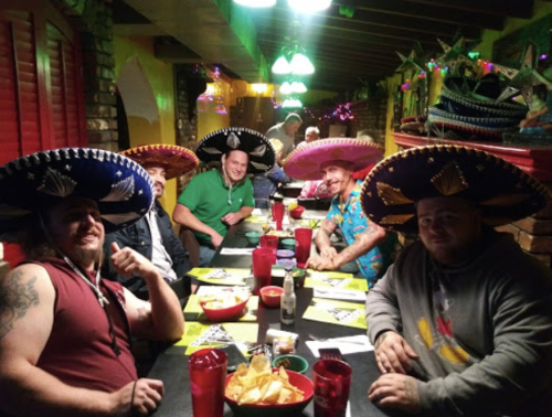 A group of friends wearing sombreros at a festive restaurant table with chips and drinks. Colorful decor in the background.