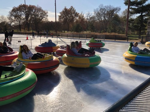 Colorful bumper cars on an ice rink, with people enjoying a fun day outdoors in a park setting.