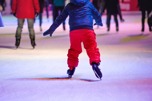 A child in red pants skates on an ice rink, surrounded by blurred figures in colorful clothing.