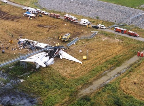 Aerial view of a plane crash site with emergency vehicles and responders on the scene in a grassy area.