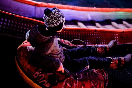A child in a winter hat sits in a colorful inner tube, ready to slide down a snowy slope.