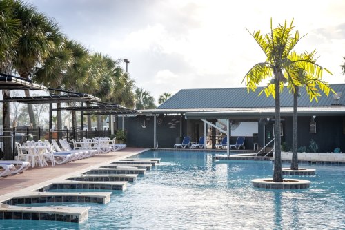 A serene pool area with lounge chairs, palm trees, and a building in the background under a bright sky.
