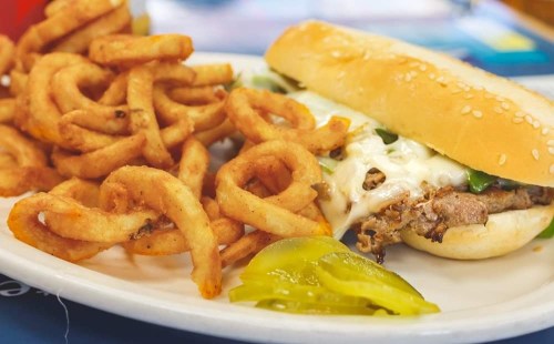A plate featuring a sandwich with meat and toppings, alongside a serving of curly fries and pickles.