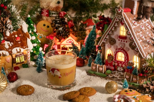 A festive scene with gingerbread houses, holiday decorations, a glass of creamy drink, and cookies on a snowy surface.