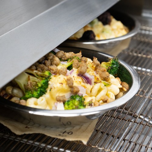 A close-up of a metal bowl filled with pasta, broccoli, and toppings, partially covered by a metal lid.