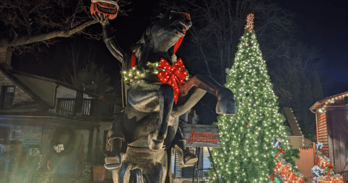 A festive statue of a horse holding a basket, adorned with a red bow, next to a brightly lit Christmas tree.