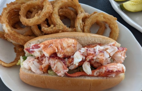 A lobster roll filled with chunks of lobster on a bun, served with crispy onion rings on the side.