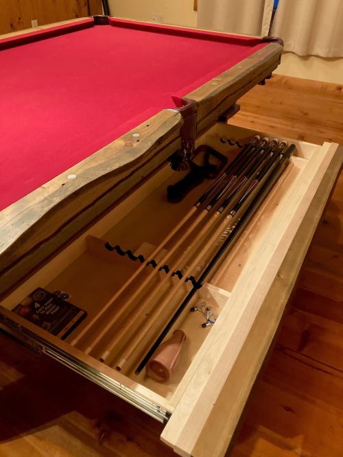 A pool table with a red felt surface and an open drawer displaying several pool cues and accessories.