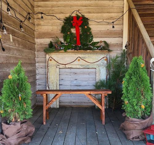 A rustic porch with a wooden bench, decorated with a wreath and greenery, flanked by two small evergreen trees.