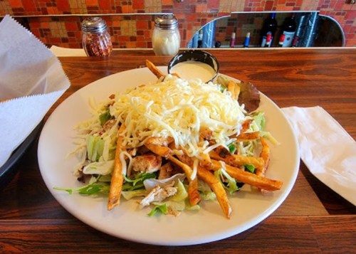 A large plate of salad topped with grilled chicken, French fries, and shredded cheese, served with a side of dressing.