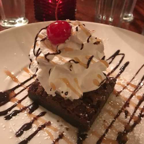 A rich brownie topped with whipped cream, chocolate drizzle, caramel, and a cherry on a white plate.