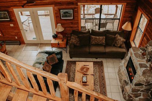 Cozy living room with a stone fireplace, wooden accents, and a view of a deck through large windows.