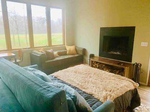 Cozy living room with a sofa, a bed, a fireplace, and large windows overlooking a green landscape.