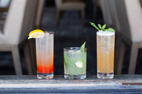 Three colorful cocktails on a wooden surface: a red drink with a lemon slice, a green mint drink, and a golden fizzy drink.