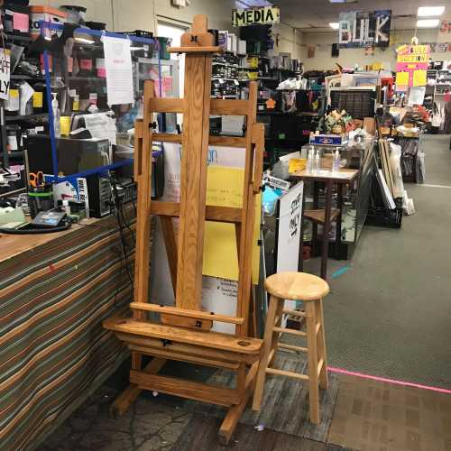 A wooden easel and a stool in a cluttered art supply store with various items in the background.