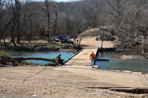 A person walks along a gravel path by a river, with vehicles parked nearby and trees in the background.