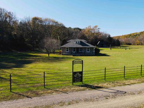 A charming house on a grassy field, surrounded by trees, with a sign at the entrance and a gravel road leading up to it.