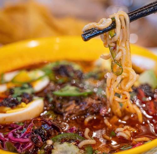 A bowl of vibrant noodle soup with vegetables, herbs, and a pair of chopsticks lifting noodles from the dish.