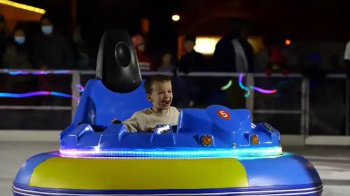 A joyful child rides a blue bumper car on an ice rink, surrounded by colorful lights and spectators in the background.