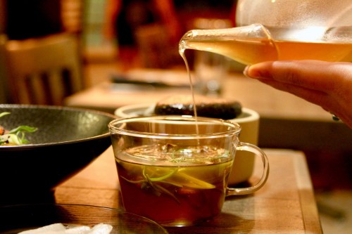 A hand pours tea from a glass teapot into a clear cup, with a blurred background of a cozy dining setting.