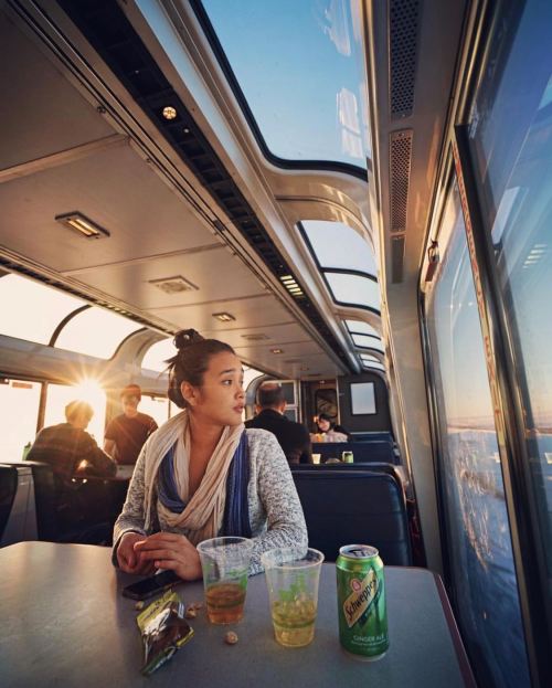 A woman sits at a table in a train, gazing out the window at a sunset, with drinks and snacks nearby.