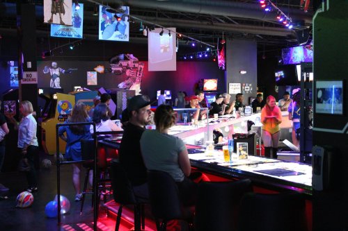 A lively bar scene with patrons at the counter, colorful lights, and arcade games in the background.