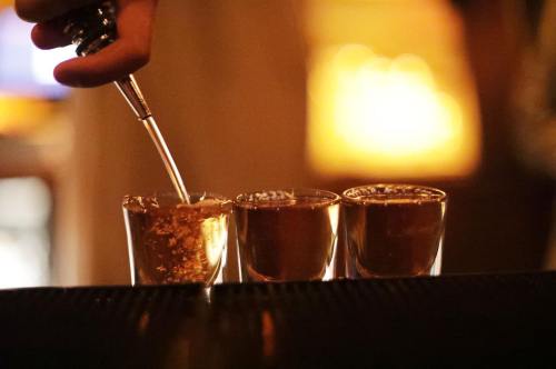 A hand pours a drink into three shot glasses on a bar, with a warm glow in the background.