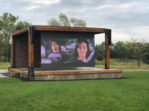 A large outdoor screen displays a scene of two people in a car, surrounded by green grass and trees.