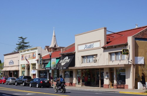 A street view of small shops with colorful storefronts and a church steeple in the background under a clear blue sky.