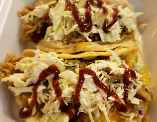 Two tacos filled with shredded meat, topped with coleslaw and drizzled with sauce, served on a white tray.