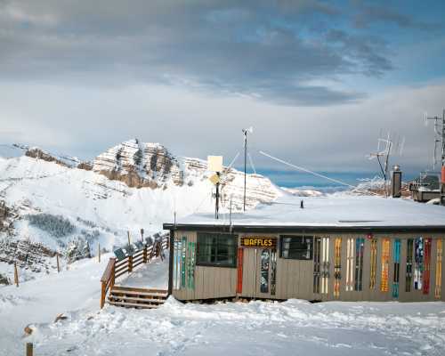 A cozy mountain hut with a "Waffles" sign, surrounded by snow-covered peaks and a cloudy sky.