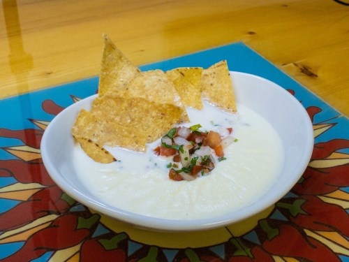 A bowl of creamy soup topped with salsa and tortilla chips, set on a colorful patterned surface.