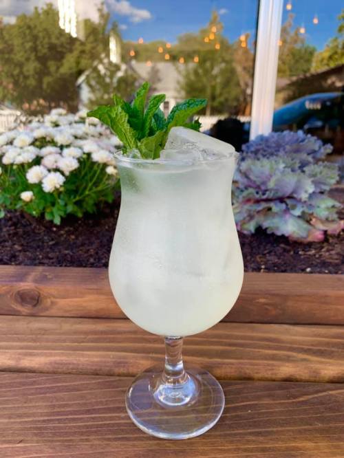 A refreshing cocktail in a glass, garnished with mint, sitting on a wooden table with flowers in the background.