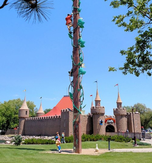A colorful playground with a castle structure, featuring children climbing a tall pole surrounded by trees and blue sky.