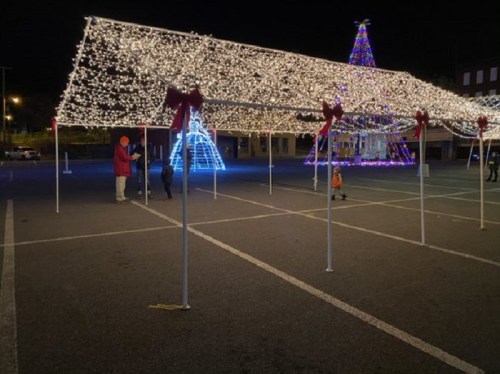 A festive display of Christmas lights in a parking lot, with decorated trees and people enjoying the scene.