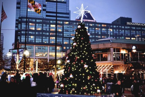 A decorated Christmas tree with lights and ornaments stands in a bustling plaza, surrounded by festive decorations and people.