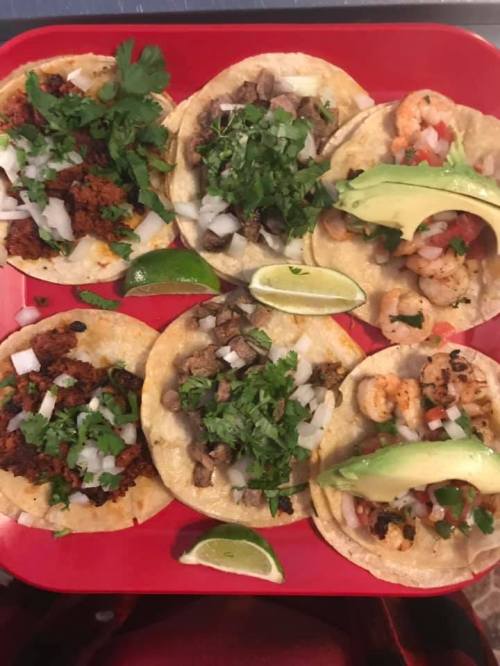 A red plate with six tacos topped with meat, shrimp, onions, cilantro, and avocado, garnished with lime wedges.
