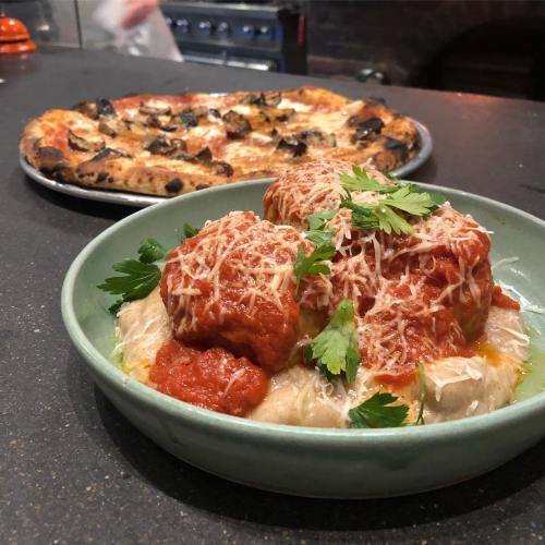 A bowl of meatballs in marinara sauce topped with cheese and parsley, with a pizza in the background.