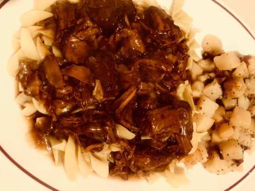 A plate of beef stew with noodles and diced potatoes, garnished with a rich brown gravy.
