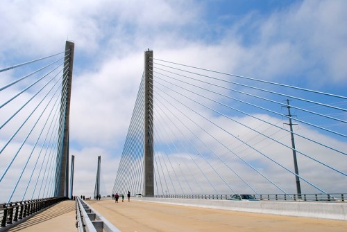 A modern cable-stayed bridge with tall towers and blue cables, under a cloudy sky, with people walking on the roadway.
