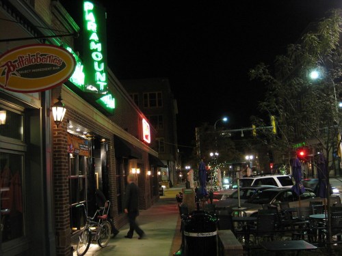 A nighttime street scene featuring a restaurant with a neon sign, outdoor seating, and parked cars.