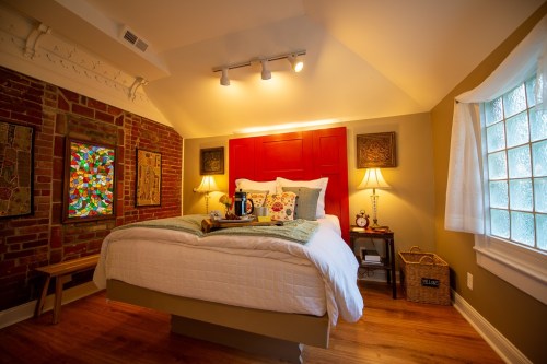 Cozy bedroom with a red headboard, decorative pillows, and a stained glass window, featuring warm lighting and wooden accents.