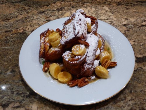 A plate of French toast topped with banana slices, pecans, and powdered sugar on a marble countertop.