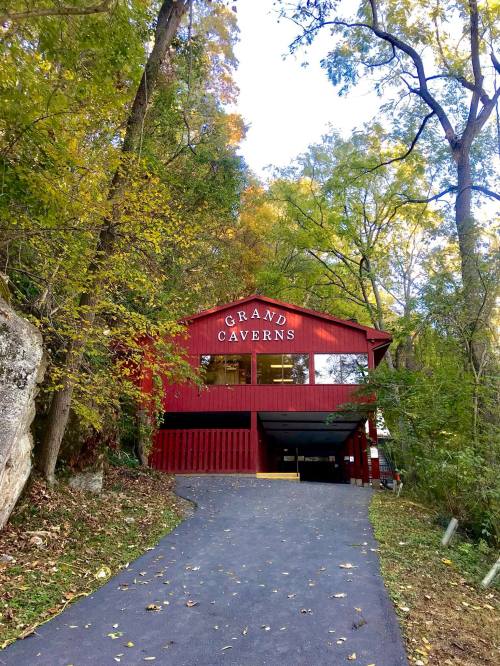 A red building labeled "Grand Caverns" surrounded by trees and foliage, with a paved path leading up to it.