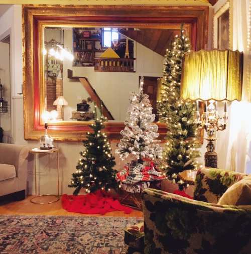 Cozy living room decorated for Christmas, featuring a large mirror, two small trees, and festive red accents.