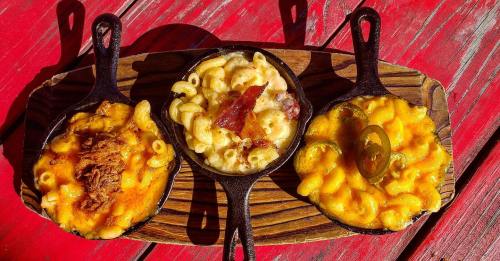 Three small cast iron skillets filled with different types of macaroni and cheese, garnished with toppings, on a wooden board.