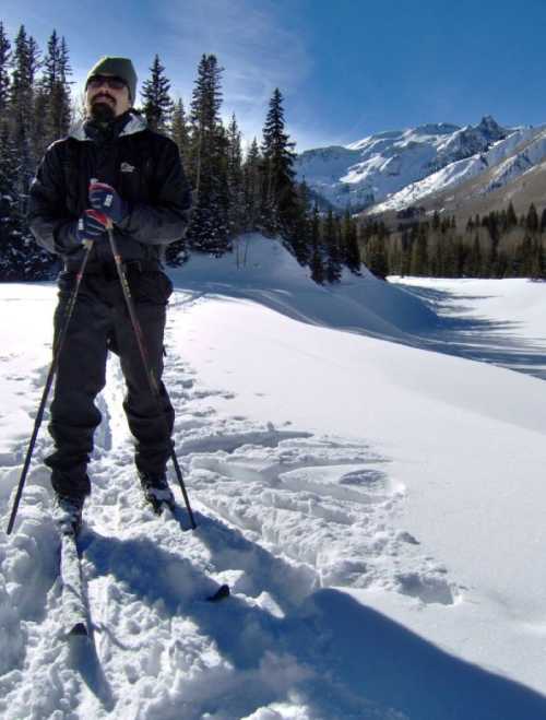 A person stands on snow-covered terrain, holding ski poles, with mountains and trees in the background under a clear blue sky.