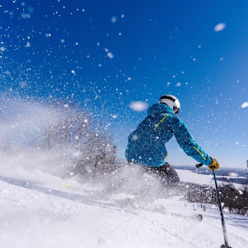 A skier in a blue jacket glides down a snowy slope under a clear blue sky, with snow flying around.